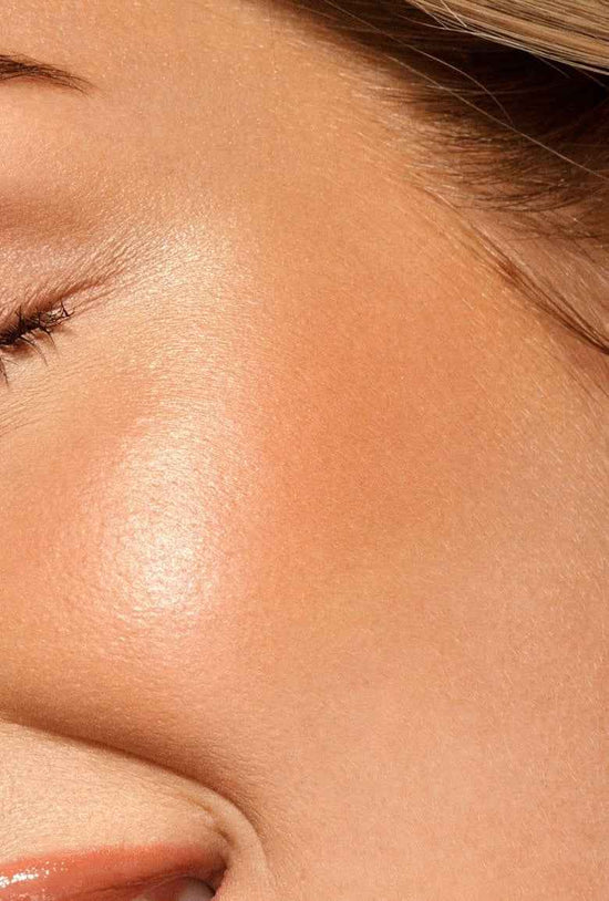 A tanned woman with peach cream blush on her cheekbones and this is a close up.