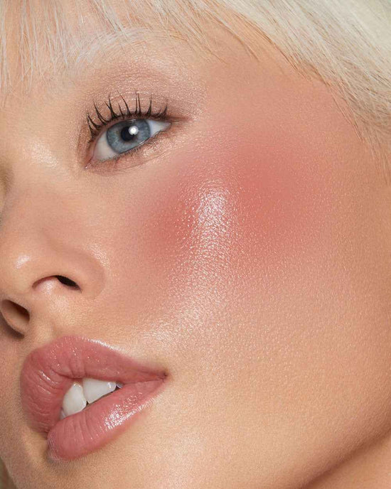 A white Caucasian woman with berry red cream blush on her cheekbones and clear lip balm gloss on her lips. This is a close up 
