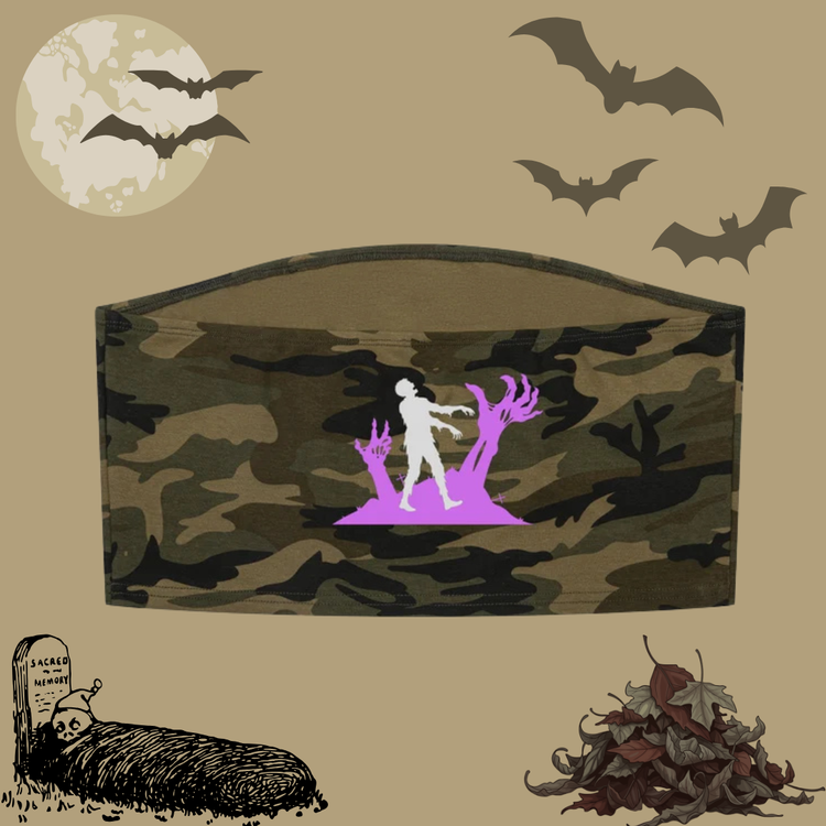A Halloween-themed decorative cropped top with a camouflage pattern and a central graphic of a white figure walking toward purple zombie hands and branches. The cropped top is displayed on a beige background decorated with bats, a full moon, a tombstone, and autumn leaves.