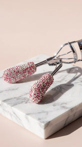A close-up of an eyelash curler embellished with pink and silver rhinestones, resting on a white marble block against a soft pink background.