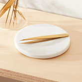 A pair of gold metal tweezers resting on a round white ceramic dish, set on a light wood surface with a glass container of similar tweezers blurred in the background.