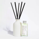 A reed diffuser set with a matte white ceramic bottle, five black reeds, and a small clear glass bottle of fragrance oil with a silver cap and a printed label, photographed on a clean white background.
