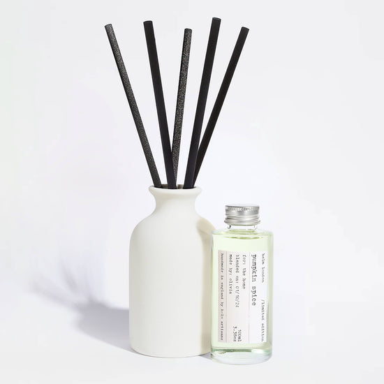 A reed diffuser set with a matte white ceramic bottle, five black reeds, and a small clear glass bottle of fragrance oil with a silver cap and a printed label, photographed on a clean white background.