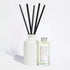 A reed diffuser set with a matte white ceramic bottle, five black reeds, and a small clear glass bottle of fragrance oil with a silver cap and a printed label, photographed on a clean white background.