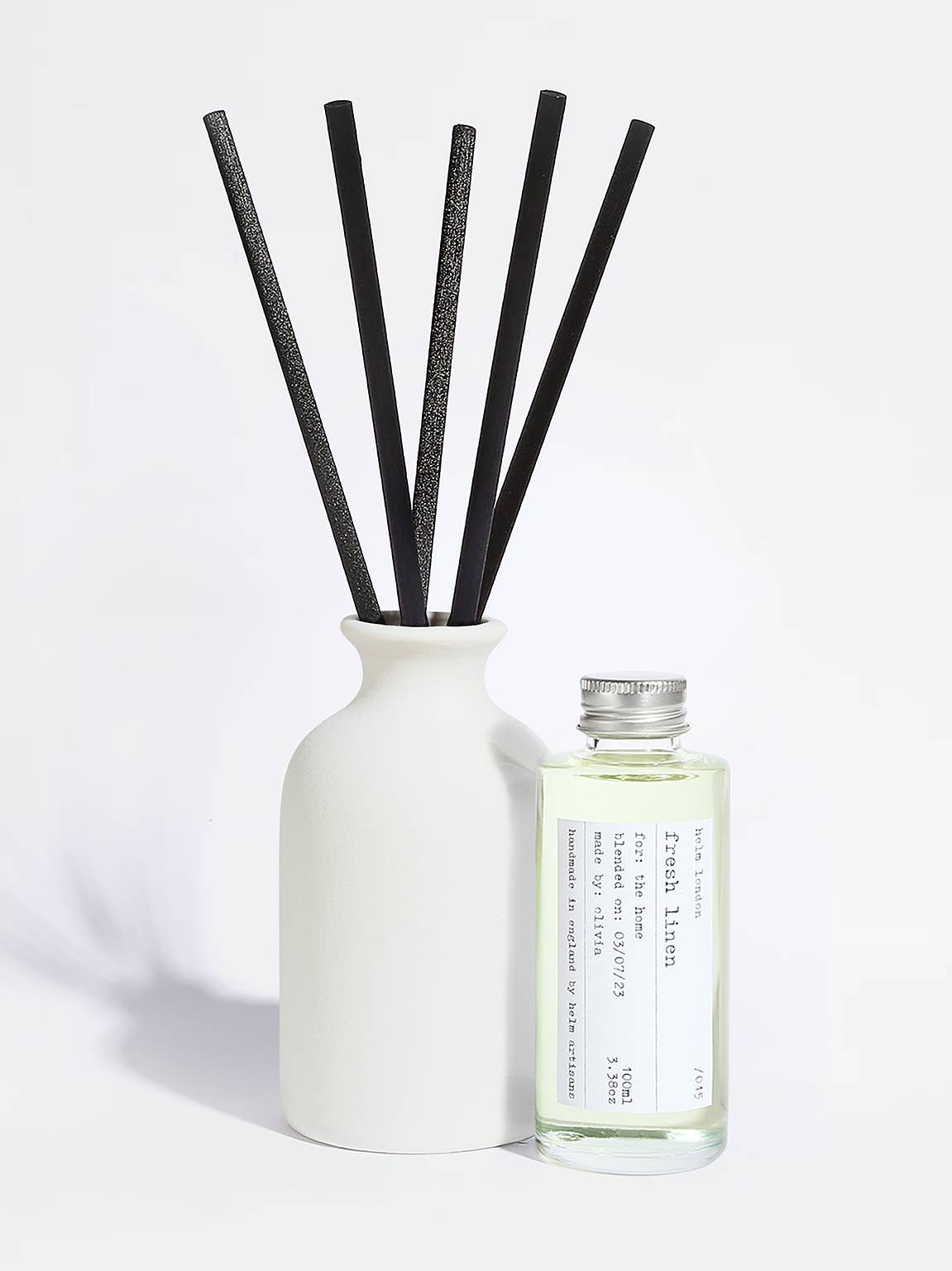 A reed diffuser set with a matte white ceramic bottle, five black reeds, and a small clear glass bottle of fragrance oil with a silver cap and a white label, photographed on a clean white background.