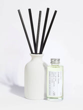 A reed diffuser set with a matte white ceramic bottle, five black reeds, and a small clear glass bottle of fragrance oil with a silver cap and a white label, photographed on a clean white background.