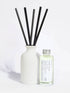 A reed diffuser set with a matte white ceramic bottle, five black reeds, and a small clear glass bottle of fragrance oil with a silver cap and a white label, photographed on a clean white background.