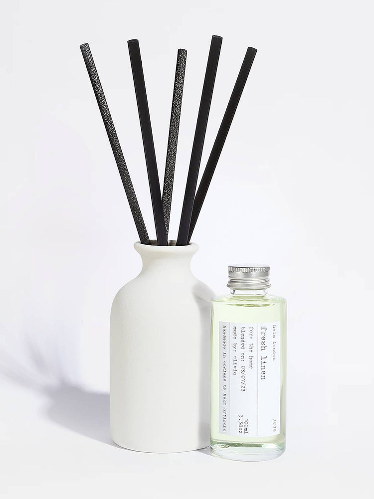 A reed diffuser set with a matte white ceramic bottle, five black reeds, and a small clear glass bottle of fragrance oil with a silver cap and a white label, photographed on a clean white background.