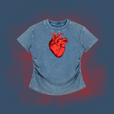 A short-sleeve t-shirt in a washed blue denim-like fabric, featuring a prominent red anatomical heart graphic on the chest, set against a dark blue background with a red glow effect.