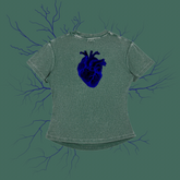 A short-sleeve t-shirt in a washed sage green color, featuring a detailed dark blue anatomical heart graphic on the chest. The shirt is displayed against a matching sage green background decorated with dark blue branching veins.