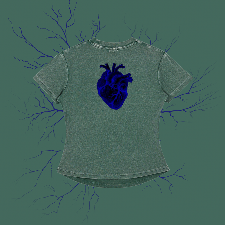A short-sleeve t-shirt in a washed sage green color, featuring a detailed dark blue anatomical heart graphic on the chest. The shirt is displayed against a matching sage green background decorated with dark blue branching veins.