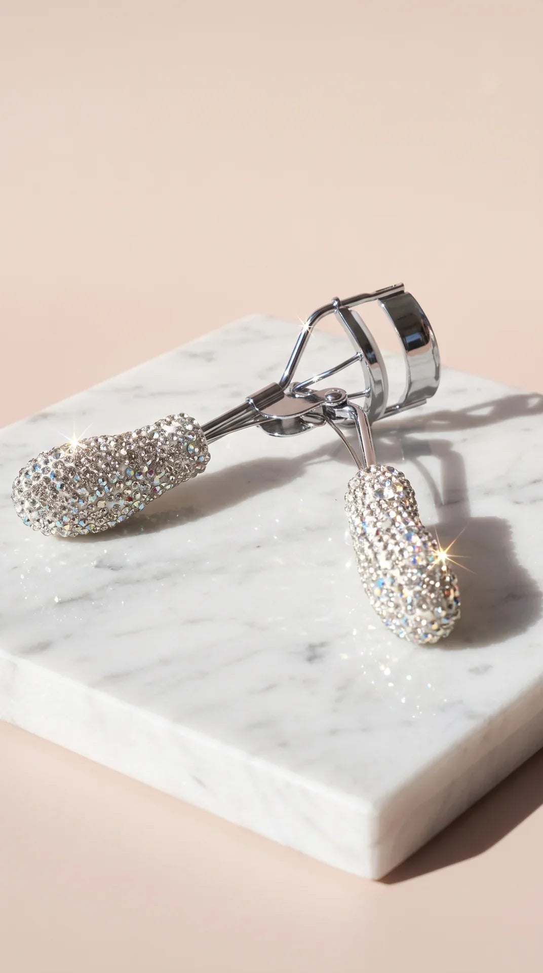 A silver eyelash curler encrusted with sparkling clear rhinestones, resting on a white marble block against a soft beige background.