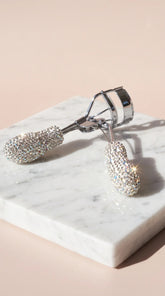 A silver eyelash curler encrusted with sparkling clear rhinestones, resting on a white marble block against a soft beige background.