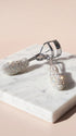 A silver eyelash curler encrusted with sparkling clear rhinestones, resting on a white marble block against a soft beige background.