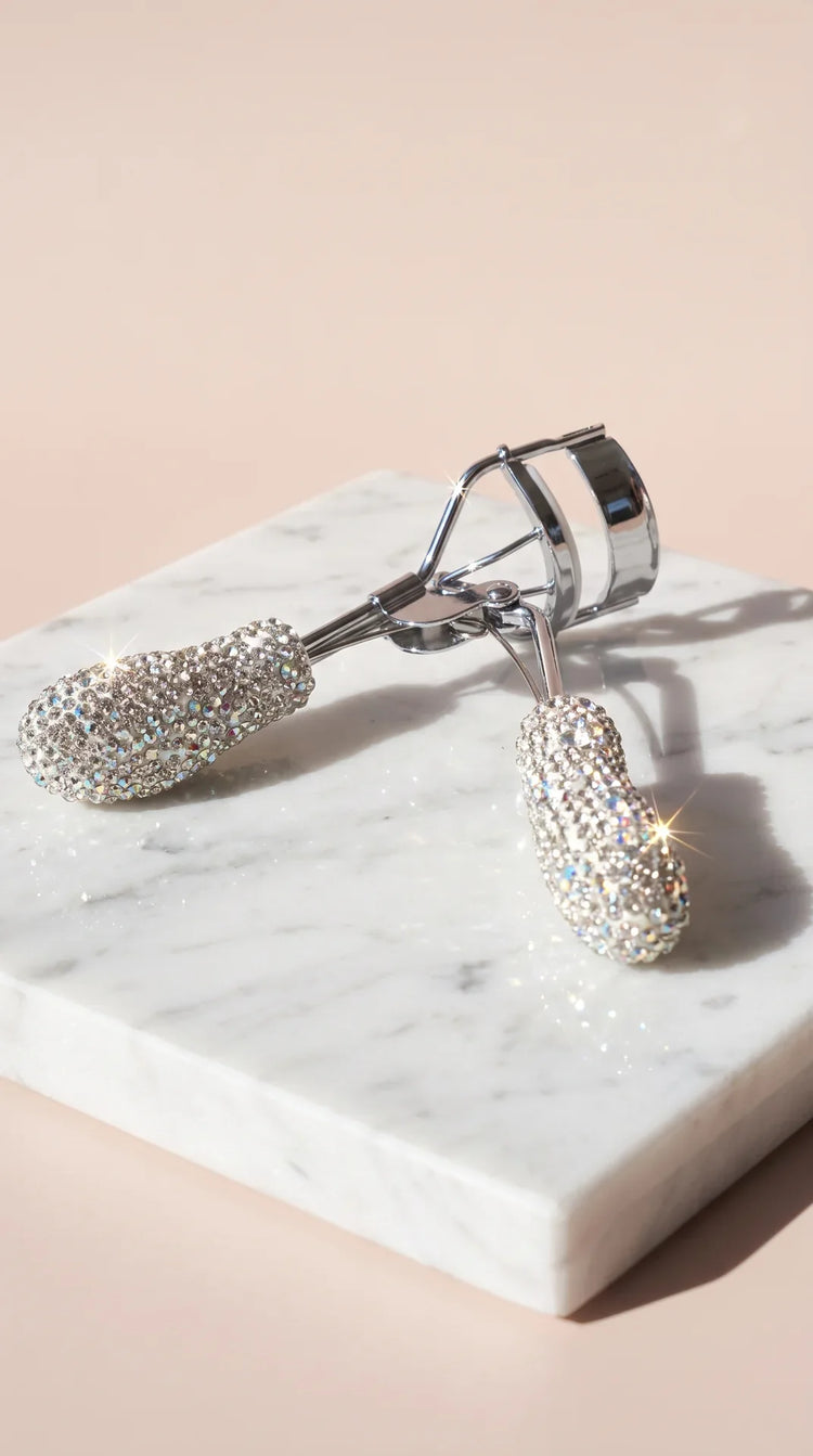 A silver eyelash curler encrusted with sparkling clear rhinestones, resting on a white marble block against a soft beige background.