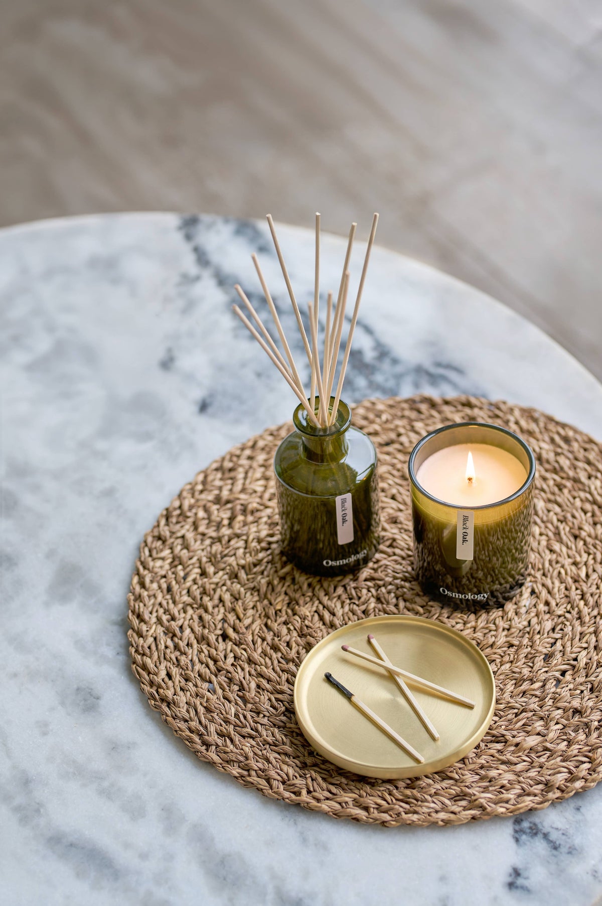 A styled home decor arrangement featuring a reed diffuser in a textured green glass bottle with wooden reeds, a lit candle in a matching green glass holder, and a small gold tray with matchsticks, all placed on a woven round placemat over a marble surface.
