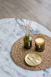 A styled home decor arrangement featuring a reed diffuser in a textured green glass bottle with wooden reeds, a lit candle in a matching green glass holder, and a small gold tray with matchsticks, all placed on a woven round placemat over a marble surface.