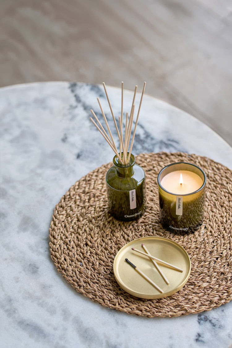 A styled home decor arrangement featuring a reed diffuser in a textured green glass bottle with wooden reeds, a lit candle in a matching green glass holder, and a small gold tray with matchsticks, all placed on a woven round placemat over a marble surface.