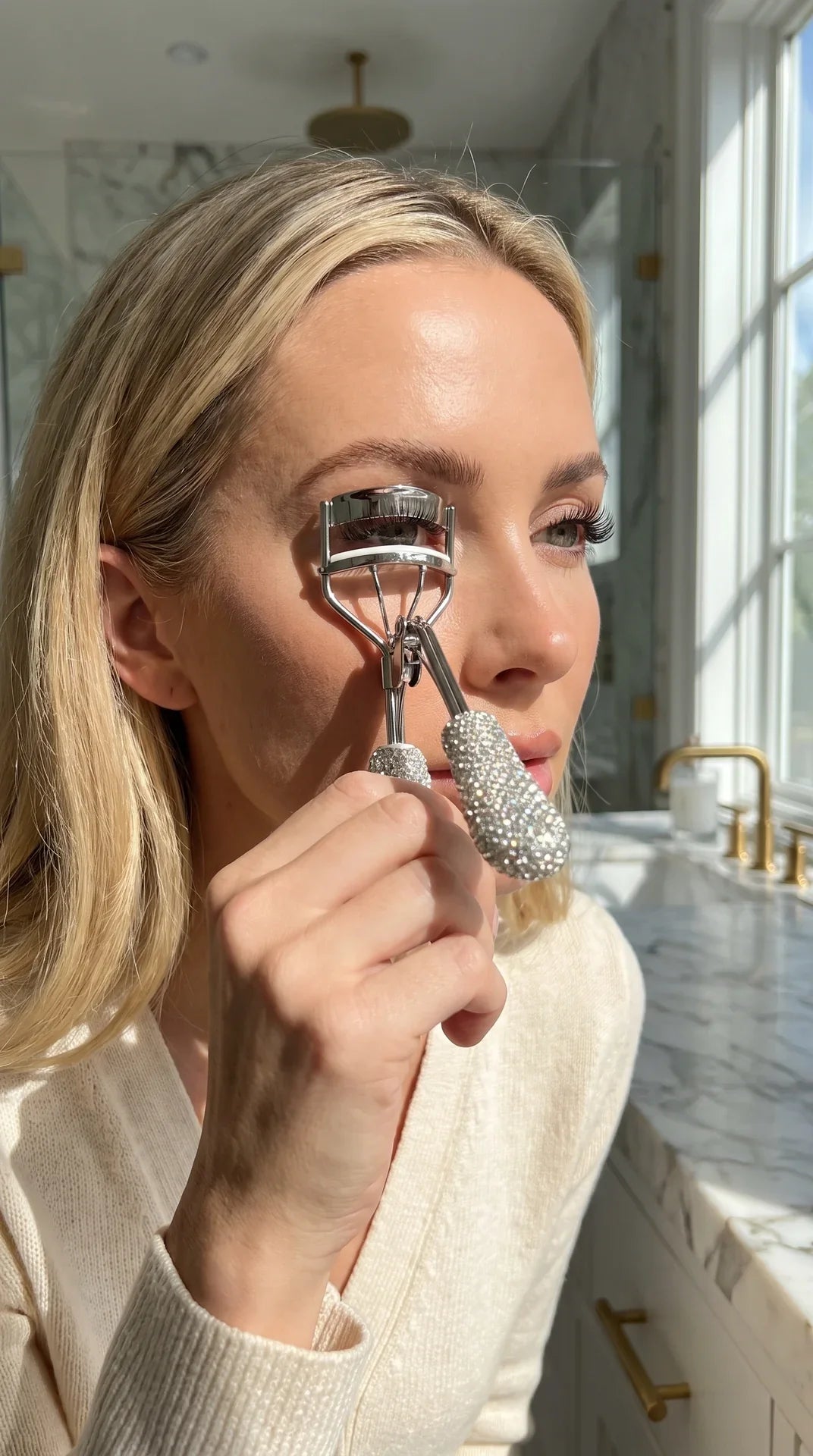 A woman in a cream knit sweater uses a crystal-embellished eyelash curler in a bright bathroom with marble countertops.