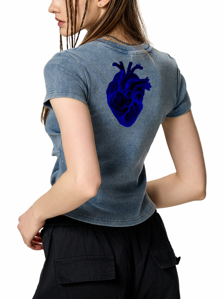 A woman shown from the back wearing a faded blue-gray cropped T-shirt with a large dark blue anatomical heart graphic on the upper back, paired with black cargo pants against a plain white background.