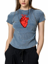 A woman wearing a blue-gray mineral-wash t-shirt with a red anatomical heart graphic on the chest, paired with black drawstring pants, against a plain white background.