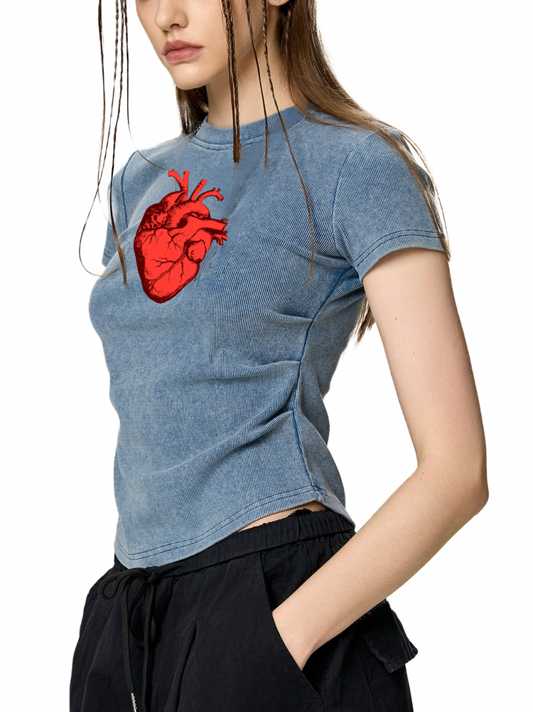 A woman wearing a cropped, heathered blue t-shirt with a red anatomical heart graphic on the chest, paired with black drawstring pants, against a plain white background.