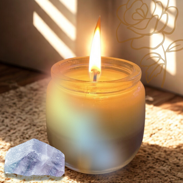 Candle in a glass jar with a lit wick, next to a rainbow fluorite crystal on a home textured surface.
