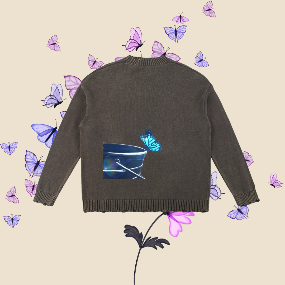 A dark grey knit sweater shown from the back, featuring a graphic print of a blue bucket with a turquoise butterfly. The sweater is displayed on a light beige background decorated with floating purple butterflies and a stylized flower.