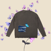 A dark grey knit sweater shown from the back, featuring a graphic print of a blue bucket with a turquoise butterfly. The sweater is displayed on a light beige background decorated with floating purple butterflies and a stylized flower.