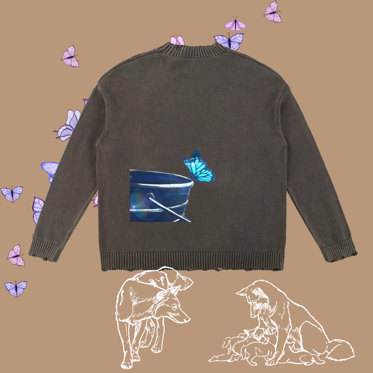 A dark grey knit sweater shown from the back, featuring a graphic print of a blue bucket with a blue butterfly and a distressed, frayed hem. The sweater is displayed on a tan background decorated with purple butterflies, line drawings of a wolf and a mother dog with her puppies, and small pink flower accents. 