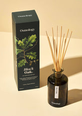 Osmology Black Oak reed diffuser set with a dark amber glass bottle, natural wood reeds, and its black packaging box featuring an oak leaf illustration, on a warm beige background.