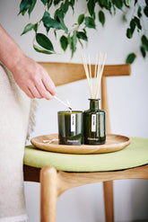 Person lighting a candle next to a diffuser on a wooden table with greenery in the background