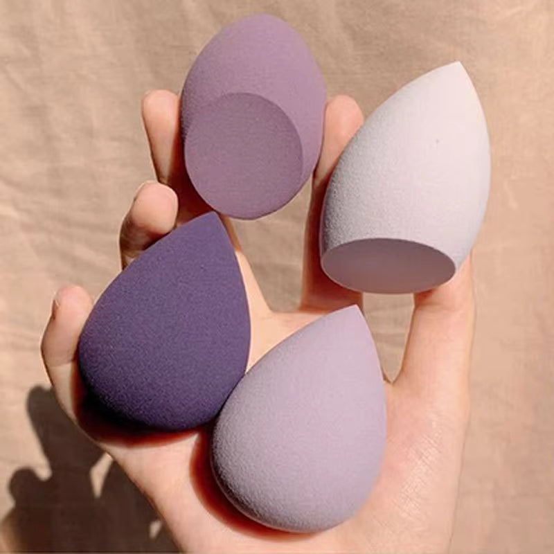 Set of four purple and grey makeup sponges held in a hand against a beige background
