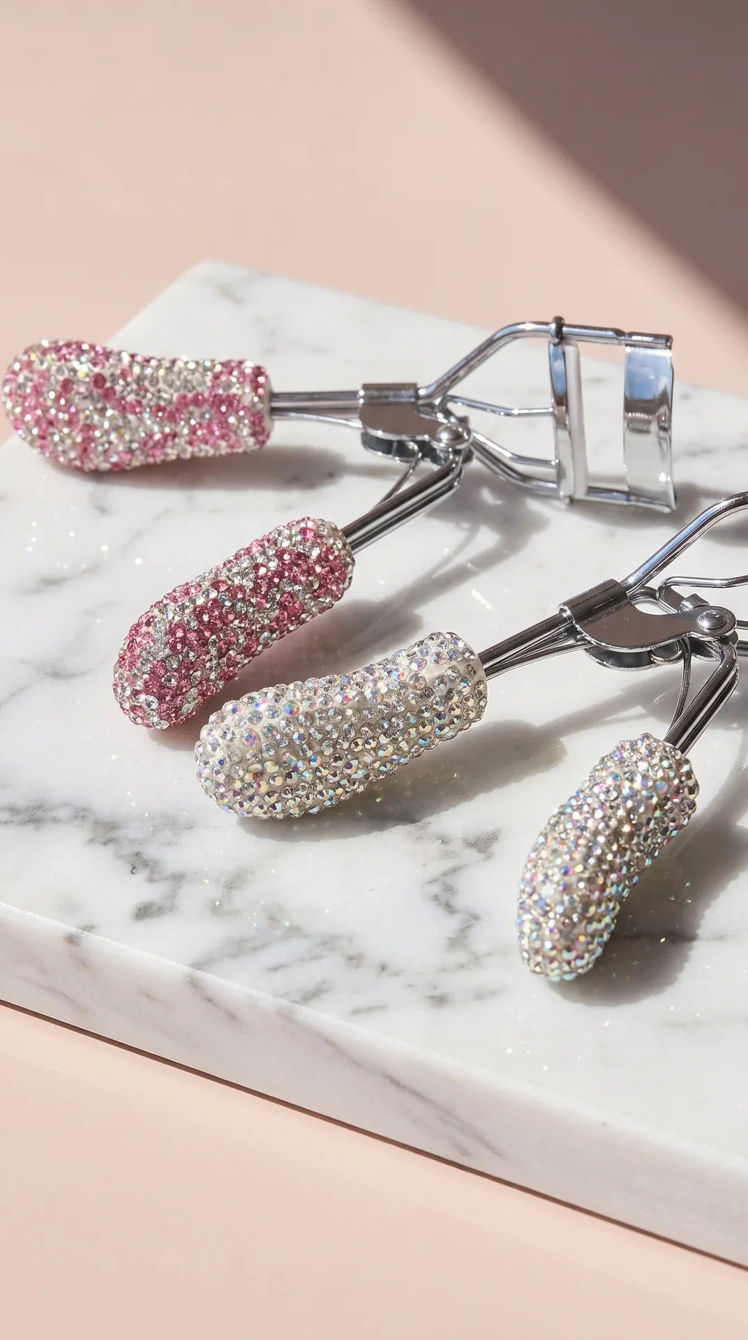 Two glitter with gemstones with eyelash curlers placed next to each other on a marble surface on a beige background.