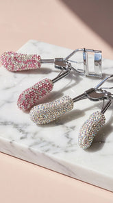 Two glitter with gemstones with eyelash curlers placed next to each other on a marble surface on a beige background.